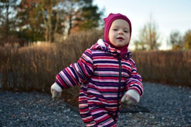 Çizgili kışlık takım elbiseli gülümseyen küçük kız çakıllı yolda dışarıda yürüyor. Soğuk mevsim ortamında neşeli bir çocukluk görüntüsü..
