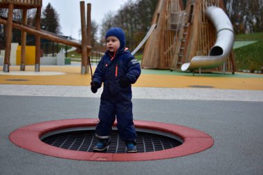 Mavi kışlık takım elbiseli çocuk elinde sopayla trambolinde oynayıp sonbahar havasının tadını çıkarıyor. Renkli modern park ve arka planda yumuşak lastik döşeme.
