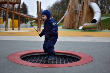 Mavi kışlık takım elbiseli çocuk elinde sopayla trambolinde oynayıp sonbahar havasının tadını çıkarıyor. Renkli modern park ve arka planda yumuşak lastik döşeme.