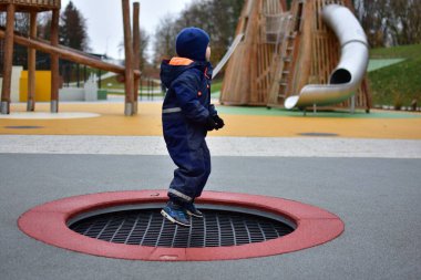 Mavi kışlık takım elbiseli çocuk elinde sopayla trambolinde oynayıp sonbahar havasının tadını çıkarıyor. Renkli modern park ve arka planda yumuşak lastik döşeme.