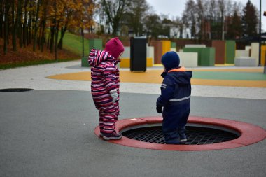 Ilık kışlık takım elbise giymiş iki çocuk renkli bir açık hava oyun alanında batık bir trambolinin yanında birlikte sonbahar havasının tadını çıkarıyorlar. Kardeşler dışarıda eğleniyor..