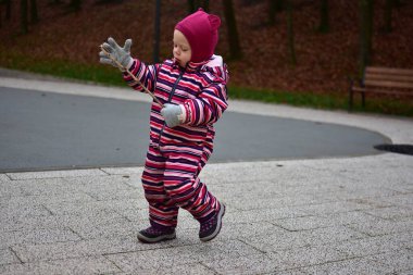 Çizgili bir kışın küçük kızı, gülerek gülerken, güz yaprakları ve park banklarıyla çevrili, modern bir açık hava oyun alanında küçük bir sopayla yürürken..