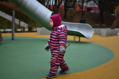 Çizgili bir kışın küçük kızı elinde küçük bir sopayla modern bir oyun parkında yürüyor. Renkli kauçuk döşeme ve eşsiz ahşap oyun yapıları arka planı dolduruyor.