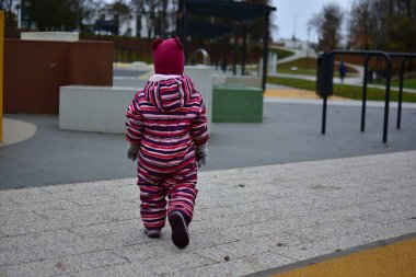 Çizgili kışın ortasında küçük bir kız modern bir açık hava oyun alanında kameradan uzaklaşıyor. Renkli geometrik şekiller ve yumuşak yüzeyler eğlenceli bir şehir ortamı yaratır.