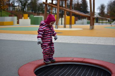 Çizgili kışın ortasında küçük bir kız batık bir trambolinin kenarında oynamaya hazırlanıyor. Renkli modern park ve yumuşak döşeme parlak bir sonbahar sahnesi yaratır.