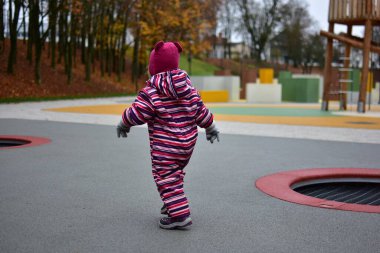 Çizgili bir kışın küçük kızı arka planda parlak lastik döşeme ve ahşap oyun yapılarıyla modern bir açık hava oyun alanında güvenle yürüyor..