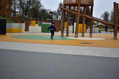 Mavi kışın ortasında küçük bir çocuk, bulutlu bir sonbahar gününde ahşap tırmanma yapıları ve yumuşak kauçuk döşemeleri olan renkli, modern bir oyun bahçesinde koşuyor..
