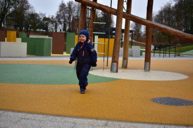 Mavi bir kış gününde gülümseyen küçük çocuk, havalı bir sonbahar gününde, ahşap tırmanma yapıları ve yumuşak kauçuk döşemeleri olan renkli, modern bir oyun parkında yürüyor..