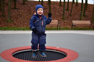Mavi bir kışın ortasında, yerdeki bir trambolinde zıplayan küçük çocuk. Arkasında park bankları, sonbahar ağaçları ve bulutlu bir günde yumuşak kauçuk döşemeler var..