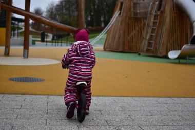 Çizgili bir kışta, arka planda tahta tırmanış yapıları ve slaytlarıyla renkli bir oyun parkında kameradan uzakta dengeli bir bisiklete binen küçük bir kız..