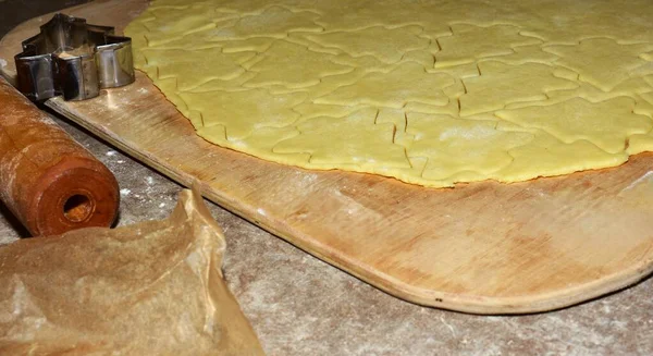 Kurabiye hamuru tahta bir tahtanın üzerine yuvarlandı ve Noel ağacı şeklinde kesildi. Yakınlarda bir metal kesici ve merdane var. Evde tatil kurabiyeleri hazırlama sürecini gösteriyor..