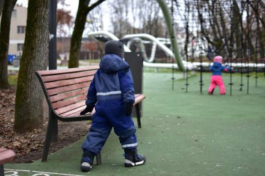 Kardeşi arka planda oynarken, çocuk parkta bir bankın yanında yürüyor. Çocuklar aile dostu park ortamında bağımsız açık hava oyunlarından hoşlanırlar..