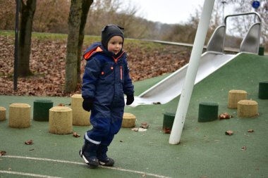 Küçük çocuk güz parkındaki kaydırağın yanında güvenle yürüyor. Kışlık giysili çocuk, soğuk sonbahar günlerinde dışarıda oyun alanlarını bağımsız olarak keşfeder..
