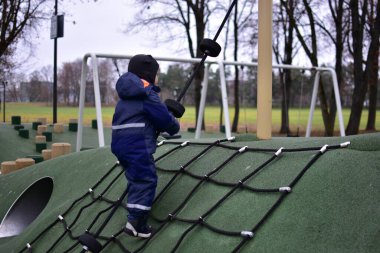 Çocuk oyun parkında ipe tırmanıyor. Çocuk, serin sonbahar havasında modern bir parkta oynarken güç, denge ve koordinasyon geliştirir..
