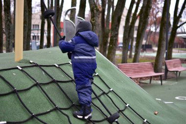 Çocuk oyun parkında ipe tırmanıyor. Çocuk, serin sonbahar havasında modern bir parkta oynarken güç, denge ve koordinasyon geliştirir..