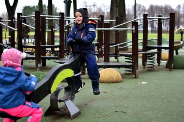 Modern bir oyun parkında oynayan iki çocuk sonbahar parkında tahterevalli gibi oynuyorlar. Sıcak kış kıyafetleri içindeki çocuklar, soğuk bulutlu bir günde açık hava etkinliğinin, dengenin ve işbirliğinin keyfini çıkarırlar..
