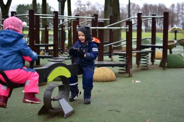 Modern bir oyun parkında oynayan iki çocuk sonbahar parkında tahterevalli gibi oynuyorlar. Sıcak kış kıyafetleri içindeki çocuklar, soğuk bulutlu bir günde açık hava etkinliğinin, dengenin ve işbirliğinin keyfini çıkarırlar..
