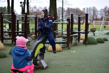 Modern bir oyun parkında oynayan iki çocuk sonbahar parkında tahterevalli gibi oynuyorlar. Sıcak kış kıyafetleri içindeki çocuklar, soğuk bulutlu bir günde açık hava etkinliğinin, dengenin ve işbirliğinin keyfini çıkarırlar..