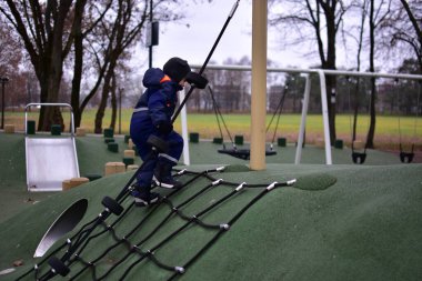 Çocuk oyun parkında ipe tırmanıyor. Çocuk, serin sonbahar havasında modern bir parkta oynarken güç, denge ve koordinasyon geliştirir..