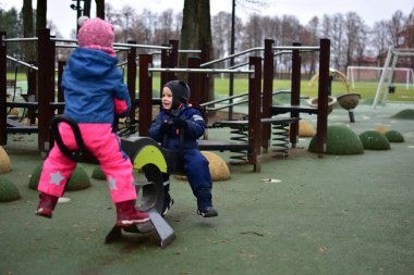 Modern bir oyun parkında oynayan iki çocuk sonbahar parkında tahterevalli gibi oynuyorlar. Sıcak kış kıyafetleri içindeki çocuklar, soğuk bulutlu bir günde açık hava etkinliğinin, dengenin ve işbirliğinin keyfini çıkarırlar..