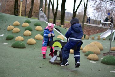 Çocuk parkında oynayan mutlu kız tepenin yanında tahterevalli gibi oynuyordu. Kışlık ceket giyen ve şapka takan çocuk, sonbaharın sonlarında parkta aktif olarak oyun oynamaktan hoşlanıyor.