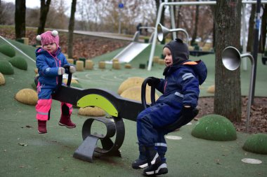 Sonbahar parkında birlikte oynayan iki çocuk tahterevalli. Sıcak kış kıyafetleri içindeki çocuklar işbirlikçi açık hava oyunları, denge ve sosyal becerileri öğrenmekten hoşlanırlar..