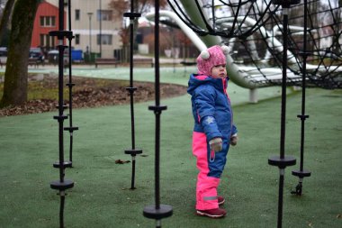 Sonbahar parkında ipe tırmanmış, parlak kışlık giysiler giymiş küçük bir kız. Modern oyun sahasında küçük çocuklar için açık hava aktivitesi, denge ve koordinasyon gelişimi.