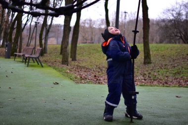 Sonbahar parkında iple oyun parkına bakan ve elinde ip tutan, kışlık giysiler giyen çocuk. Açık hava oyunu dengeyi, koordinasyonu ve motor becerileri geliştirmeyi destekler.