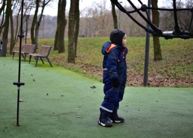 Küçük çocuk, sonbahar boyunca dışarıda oyun alanında halat tırmanışı yapısının yanında duruyor. Sıcak kış giysileri giyen çocuk oyun parkı ekipmanlarını keşfediyor, parktaki erken çocukluk gelişimi.