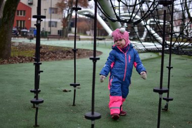 Sonbahar parkında ipe tırmanmış, parlak kışlık giysiler giymiş küçük bir kız. Modern oyun sahasında küçük çocuklar için açık hava aktivitesi, denge ve koordinasyon gelişimi.