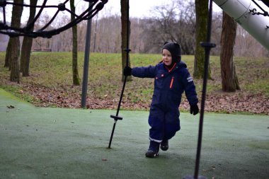 Küçük çocuk, sonbahar boyunca dışarıda oyun alanında halat tırmanışı yapısının yanında duruyor. Sıcak kış giysileri giyen çocuk oyun parkı ekipmanlarını keşfediyor, parktaki erken çocukluk gelişimi.