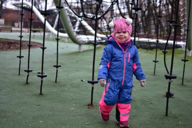 Sonbahar parkında ipe tırmanmış, parlak kışlık giysiler giymiş küçük bir kız. Modern oyun sahasında küçük çocuklar için açık hava aktivitesi, denge ve koordinasyon gelişimi.