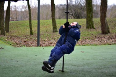 Sonbahar parkında iple oyun oynayan küçük bir çocuk. Sıcak kış giysileri giyen çocuk açık hava oyunlarının keyfini çıkarıyor, dengeyi, koordinasyonu ve motor becerileri güçlendiriyor.