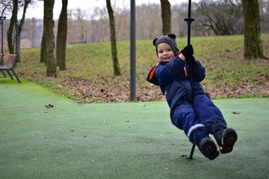 Sonbahar parkında iple oyun oynayan küçük bir çocuk. Sıcak kış giysileri giyen çocuk açık hava oyunlarının keyfini çıkarıyor, dengeyi, koordinasyonu ve motor becerileri güçlendiriyor.