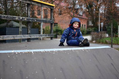 Sonbahar boyunca oyun parkında kaykay rampasının tepesinde oturan mutlu çocuk. Sıcak kışlık giysiler giyen çocuk, açık hava oyunlarından zevk alıyor, erken çocuklukta güven, denge ve koordinasyon geliştiriyor..