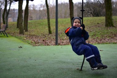 Sonbahar parkında iple oyun oynayan küçük bir çocuk. Sıcak kış giysileri giyen çocuk açık hava oyunlarının keyfini çıkarıyor, dengeyi, koordinasyonu ve motor becerileri güçlendiriyor.