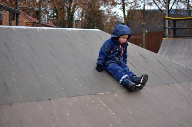 Sonbaharda çocuk oyun parkında kaykay rampasından kayıyor. Sıcak kış kıyafetleri giyen, denge ve koordinasyon uygulayan, aktif açık hava oyunlarından ve erken çocukluk keşiflerinden hoşlanan bir çocuk..