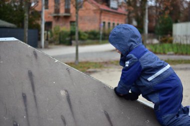 Küçük çocuk sonbahar boyunca oyun parkında kaykay rampasına tırmanıyor. Kışlık giysiler giyen çocuk aktif açık hava oyunları ve erken çocukluk eğitimi yoluyla güç, denge ve koordinasyon geliştiriyor.
