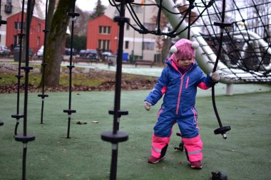 Sonbahar parkında ipe tırmanmış, parlak kışlık giysiler giymiş küçük bir kız. Modern oyun sahasında küçük çocuklar için açık hava aktivitesi, denge ve koordinasyon gelişimi.