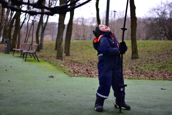 Sonbahar parkında iple oyun parkına bakan ve elinde ip tutan, kışlık giysiler giyen çocuk. Açık hava oyunu dengeyi, koordinasyonu ve motor becerileri geliştirmeyi destekler.