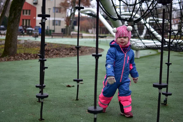 Sonbahar parkında halatla yürüyen, parka tırmanan, renkli kışlık giysiler giyen küçük bir kız. Modern oyun sahasında küçük çocuklar için açık hava aktivitesi, denge ve koordinasyon gelişimi.