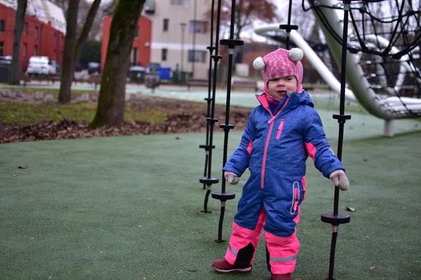 Sonbahar parkında ipe tırmanmış, parlak kışlık giysiler giymiş küçük bir kız. Modern oyun sahasında küçük çocuklar için açık hava aktivitesi, denge ve koordinasyon gelişimi.