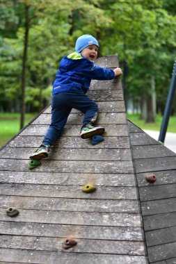 Gülen çocuk renkli tutuş ile tahta bir oyun parkı duvarına tırmanıyor. Mutlu çocuk dışarıda oynamaktan hoşlanıyor, yeşil park ortamında motor becerileri, kendine güveni ve fiziksel gücü geliştiriyor..