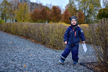 Sonbahar parkında çakıl yolda yürüyen gülümseyen çocuk. Açık hava tulumu ve şapka giyen çocuk, arka planda renkli ağaçlar ve çalılarla sonbahar sezonunun tadını çıkarıyor..
