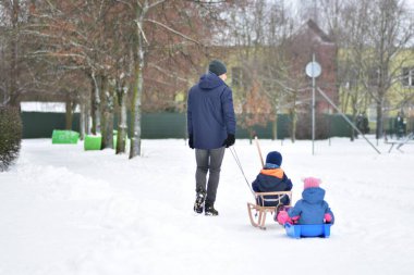 Babam, kızaktaki iki çocuğu çekerken karlı parkta yürüyor. Huzurlu bir kış aile anı, açık havada ebeveynlik, soğuk doğal ortamda çocukluk anıları..