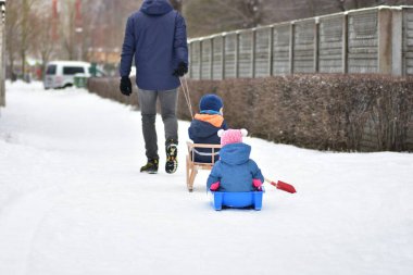 Babam kış boyunca iki küçük çocuğu kızakla çekerken karlı kaldırımda yürüyordu. Aile aktivitesi, ebeveynlik yaşam tarzı, soğuk şehir ortamında çocukluk eğlencesi ve güvenlik kavramı.