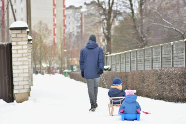 Babam kış boyunca iki küçük çocuğu kızakla çekerken karlı kaldırımda yürüyordu. Aile aktivitesi, ebeveynlik yaşam tarzı, soğuk şehir ortamında çocukluk eğlencesi ve güvenlik kavramı.