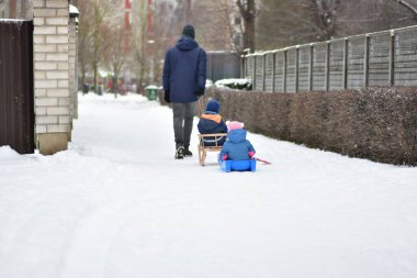 Babam kış boyunca iki küçük çocuğu kızakla çekerken karlı kaldırımda yürüyordu. Aile aktivitesi, ebeveynlik yaşam tarzı, soğuk şehir ortamında çocukluk eğlencesi ve güvenlik kavramı.