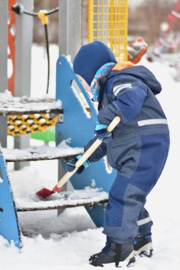 Kışın giydiği kıyafetlerle çocuk parkının merdivenlerine kar kürüyen bir çocuğun yakın çekimi. Tatlı çocukluk anı, açık hava kış aktivitesi, bağımsızlık, motor beceriler ve günlük oyun yoluyla öğrenme.