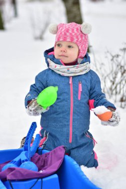 Karda duran küçük kız elinde karla dolu renkli plastik bardaklar tutuyor. Sıcak kış kıyafetleri giymiş bir çocuk açık hava oyunlarının, kış aktivitelerinin ve huzurlu çocukluk anlarının tadını çıkarıyor..